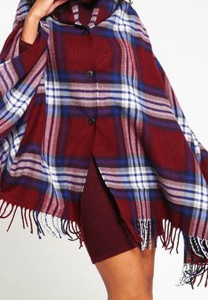 Frau trägt einen rot-blau-weiß karierten, geknöpften Poncho mit Fransen über einem kastanienbraunen Kleid und posiert mit einer Hand nahe am Hals.