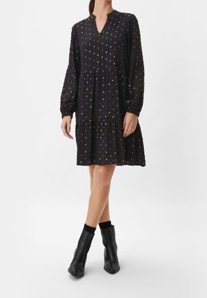 Woman wearing a black long-sleeve dress with multicolored dots, paired with black ankle boots and black socks, standing with legs crossed.