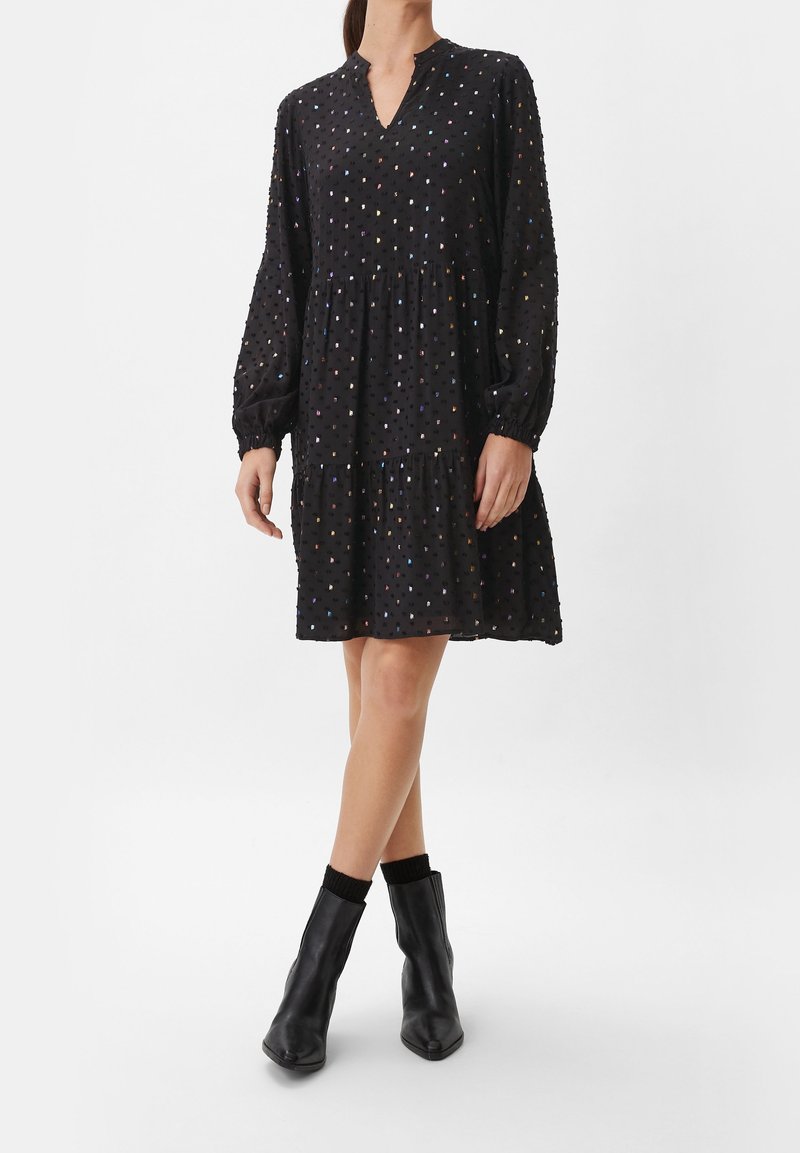 Femme portant une robe noire à manches longues avec des pois multicolores, associée à des bottines noires et des chaussettes noires, debout jambes croisées.