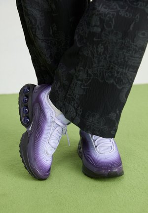 Person iført sorte bukser med blomsterprint og lilla gradient sneakers med gennemsigtige boble-såler, stående på en lysegrøn gulv.