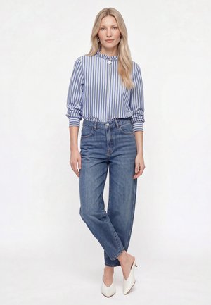 Frau mit langen blonden Haaren, die eine blau-weiß gestreifte Bluse, hoch taillierte blaue Jeans und weiße spitze Schuhe trägt, steht mit überkreuzten Beinen.