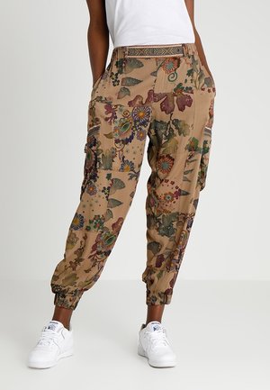 Persona con pantalones cargo beige con estampado floral, puños elásticos y bolsillos, combinados con zapatillas blancas y una camiseta blanca.