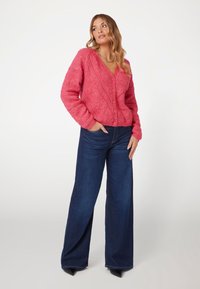 Cardigan rosa in maglia con scollatura a V, motivo testurizzato e bottoni frontali, abbinato a jeans a vita alta di colore blu scuro con gamba ampia.