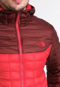 Giacca isolante rossa e marrone con texture trapuntata, colletto alto e chiusura con zip. Presenta il logo sul lato sinistro superiore.