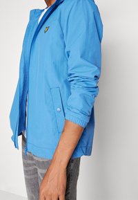 Light blue zip-up jacket with a hood, featuring a front pocket and elastic cuffs. A small black logo is visible on the chest area.