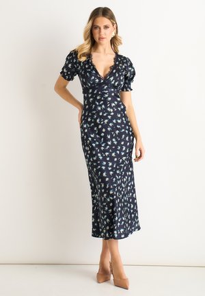 Woman posing in a navy blue floral midi dress with puff sleeves and beige heels against a plain light background.