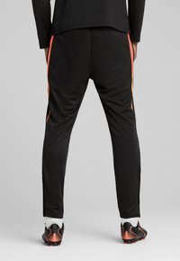 Calças atléticas pretas com riscas laterais laranja, feitas de um material sintético suave. Inclui fechos junto aos punhos.