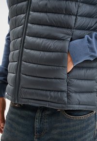 Wattierte Weste in Dunkelblau mit zipbarem Frontverschluss, gestepptem Muster in horizontalen Abschnitten und einem blauen Langarmshirt darunter.