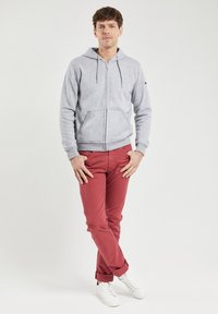Un sweat à capuche zippé gris avec des poches sur le devant, associé à un pantalon rouge et des baskets blanches. Le tissu semble doux, et les pantalons sont retroussés aux poignets.