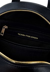 Unutrašnjost crne kožne torbice s zlatnim patentnim zatvaračem, koja sadrži jednu zip-zipnu džep i utisnuti zlatni logo "Victoria Hyde London." Glatka tekstura.