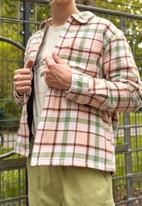 Flannel shirt in pink, green, and brown plaid with a button-up design. Features a pointed collar and chest pocket, worn over a light tee.