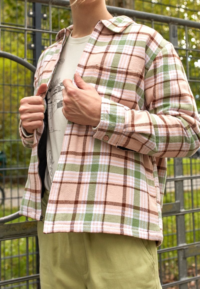 Flannel shirt in pink, green, and brown plaid with a button-up design. Features a pointed collar and chest pocket, worn over a light tee.