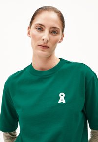 Grünes Baumwoll-T-Shirt mit rundem Ausschnitt und einem kleinen, weißen, bandförmigen Logo auf der linken Brustseite. Kurze Ärmel, glatte Textur.