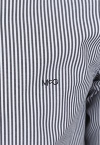 Gestreepte stof met smalle verticale lijnen in zwart en wit, met een geborduurd "McG" logo in zwart aan de linkerkant.