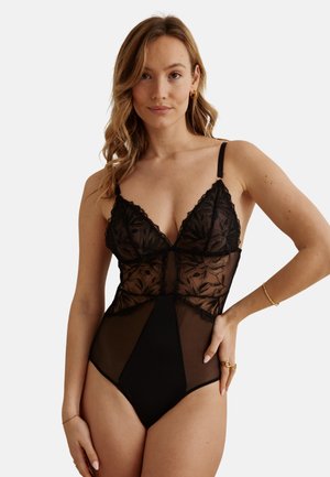 Femme portant un body noir en dentelle et résille avec des motifs floraux, debout avec une main sur la hanche, sur un fond uni.