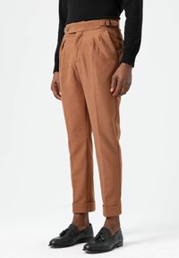 Beige broek met rechte pijpen, geplooide voorkant, zijgespsdetail, opgerolde zomen, gecombineerd met zwarte loafers op een lichte achtergrond.