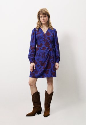 Wikkeljurk in blauw met bordeauxrode bloemenpatronen, lange mouwen en een gestrikt middel. Gepaard met bruine suède cowboylaarzen.
