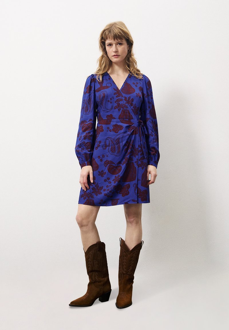 Wickelkleid in Blau mit burgunderfarbenen Blumenmustern, langen Ärmeln und gebundenem Taillengürtel. Kombiniert mit braunen Wildleder-Cowboy-Stiefeln.