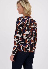 Vrouw met kort blond haar, gekleed in een langemouwen top met abstracte zwarte, witte, roestkleurige en marineblauwe bladdessins, gecombineerd met donkere spijkerbroek.