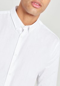 Chemise blanche à bouton avec un col classique, présentant un tissu texturé et un éclat subtil. Les boutons sont de couleur blanche assortie, créant un design minimaliste.