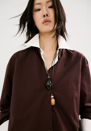 Tânără cu cămașă supradimensionată maro închis și guler alb, purtând un colier cu pandantiv negru și maro, pe un fundal simplu.