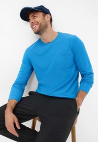 Langarmige blauwe shirt met een borstzak, gecombineerd met donkergrijze broeken. Model met een marineblauwe pet, zittend op een houten kruk.
