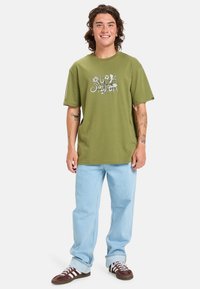 Olivově zelené bavlněné tričko s grafickým potiskem, ve spojení se světle modrými džíny s ohrnutými nohavicemi a hnědými teniskami s bílými pruhy.