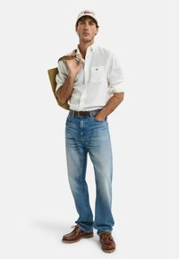 Leichtes weißes Hemd, blaue Jeans, braune Lederschuhe und eine hellbraune Tragetasche; trägt eine beigefarbene Kappe mit Logodetails.