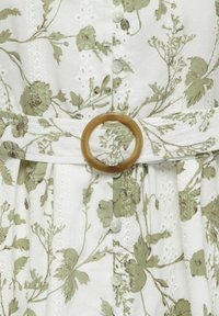 Robe en tissu blanc avec broderie florale verte, bandes de dentelle verticales, boutons sur le devant et ceinture assortie avec une boucle ronde marron.