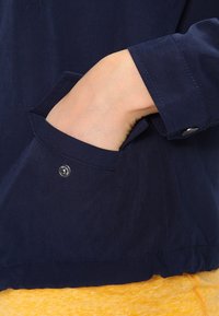 Marinblå jacka med en sidficka som har en silverfärgad tryckknapp och en slät tygtextur. Nederkanten är samlad för extra form.