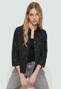 Zwarte shirt met witte hartpatronen, met knopen, lange mouwen, gecombineerd met een zwart tanktop en grijze denim broek.