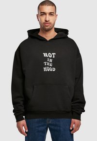 Černá mikina s kapucí a přední kapsou, s bílým textem "NOT IN THE MOOD" ve stylizovaném písmu. Vyrobena z měkké látky a s volným střihem.