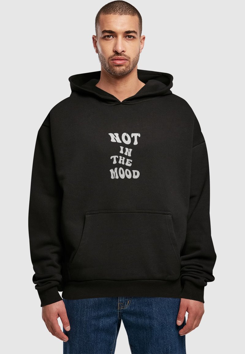 Černá mikina s kapucí a přední kapsou, s bílým textem "NOT IN THE MOOD" ve stylizovaném písmu. Vyrobena z měkké látky a s volným střihem.