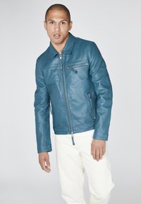 Giubbotto in pelle verde acqua con colletto, chiusura frontale con zip e due tasche con zip. Indossato sopra una camicia bianca e abbinato a pantaloni chiari.