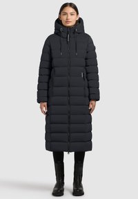 Lange zwarte pufferjas met capuchon, horizontaal doorgestikt ontwerp, ritssluiting aan de voorkant, zijzakken en zwarte enkellaarzen. Glad, waterafstotend materiaal.