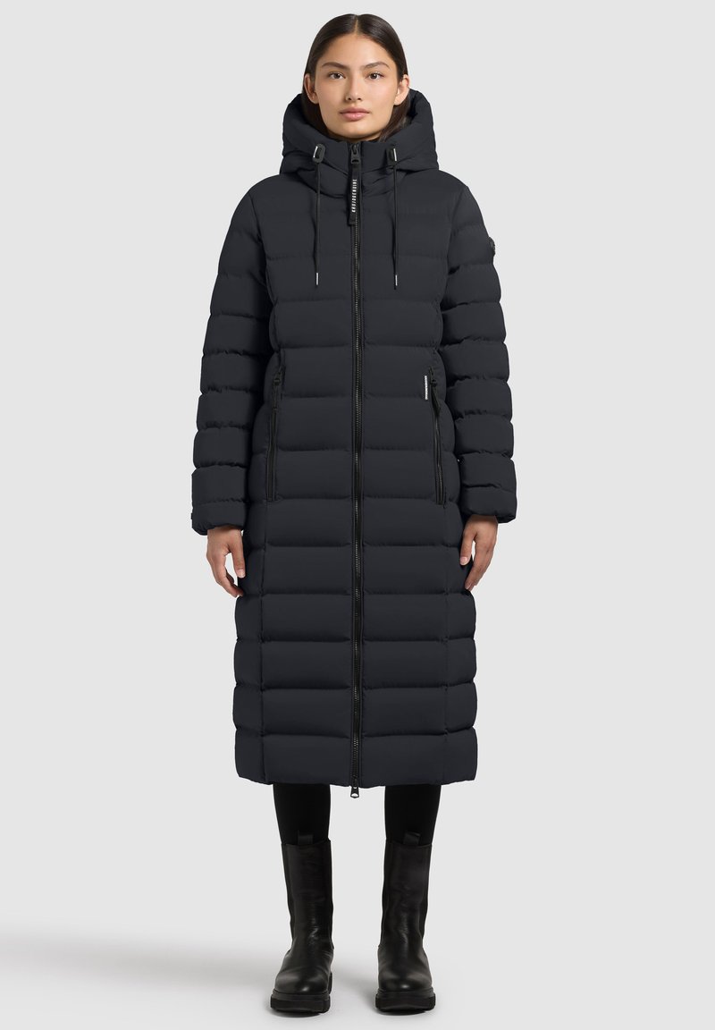 Lange zwarte pufferjas met capuchon, horizontaal doorgestikt ontwerp, ritssluiting aan de voorkant, zijzakken en zwarte enkellaarzen. Glad, waterafstotend materiaal.