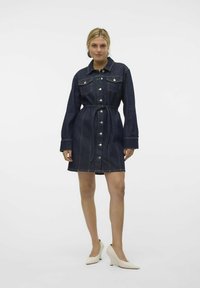 Jeans haljina u tamnoplavoj boji, s gumbima sprijeda, dvije džepove na prsima, dugim rukavima i strukiranjem s remenom. Glatka tekstura tkanine.