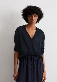 Cardigan bleu marine à manches longues avec un décolleté en V profond, fermeture par boutons à l'avant et manches retroussées, associé à une jupe taille assortie.
