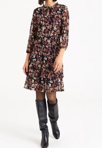 Robe noire à fleurs avec des volants superposés, présentant des manches longues, un décolleté en forme de trou de serrure et des motifs floraux multicolores. Associée à des bottes noires jusqu'à la cuisse.
