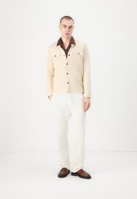 Beige gombos ing két mellzsebbel egy barna galléros ing felett, fehér nadrággal és barna cipővel párosítva, egy sima háttér előtt állva.