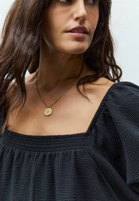 Zwarte blouse met textuur en pofmouwen, met een vierkante halslijn. Een gouden hangerketting valt op de halslijn en voegt een metalen accent toe.