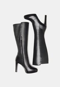 Bottes noires en cuir montantes au design élégant, avec une fermeture éclair sur le côté, un talon haut et une texture lisse. Les accents incluent une étiquette de marque.