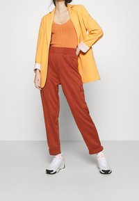 Orange ribbad linne, gul kavaj, rostfärgade cargobyxor och vita sneakers med svarta detaljer. Släta material och avslappnad passform.