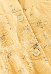 Geltona tinklelio medžiaga su mažais gėlių raštais, dvi apvalios sagos ir raukiniai palei siūlę. Tekstūruota ir lengvos išvaizdos.