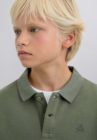 Polo de algodón verde oliva con dos botones, tela texturizada y un logo de calavera tonal en el lado izquierdo del pecho, superpuesto a una camisa blanca.
