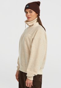 Beige fleece pullover met hoge kraag en rits aan de voorkant, met ribgebreide manchetten en onderrand, gecombineerd met een bruine muts.