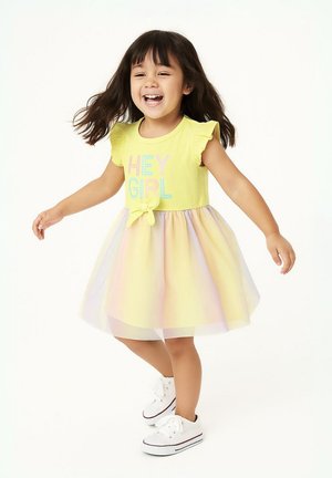 Robe jaune avec des manches courtes à volants, ornée du texte pastel "HEY GIRL". La jupe présente un dégradé allant du jaune à des teintes douces de l'arc-en-ciel. Baskets blanches.