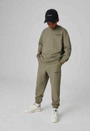 Olivgrön sweatshirt och joggers med uppvikta fållar, med svart "ALLSAINTS" logotyp, tillsammans med vita sneakers och en svart keps.