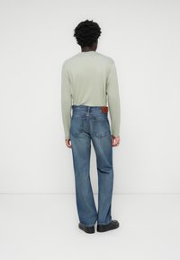 Lichtgroene blouse met lange mouwen, gecombineerd met blauwe denim jeans met een relaxte pasvorm, voorzien van een bruine leren merkpatch op de achterkant van de tailleband.
