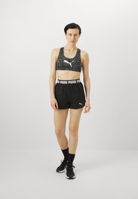 Μαύρο και γκρι καμουφλάζ αθλητικό σουτιέν με λευκό λογότυπο, συνδυασμένο με μαύρες βερμούδες που διαθέτουν λευκή επωνυμία στη μέση και μαύρα αθλητικά παπούτσια.